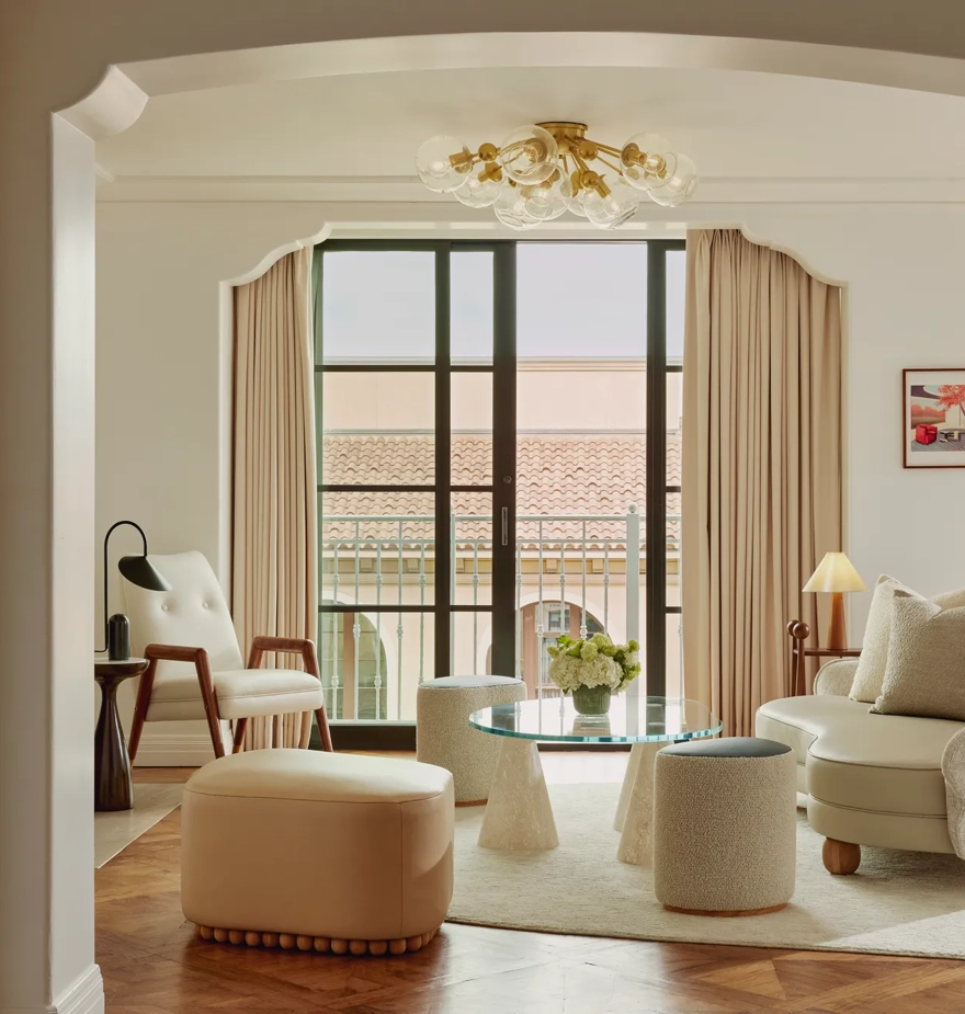 Intimate sitting room facing balcony doors; glass coffee table on conical bases, boucle stools, mid‑century chair and loveseat; terracotta roofline visible outside.