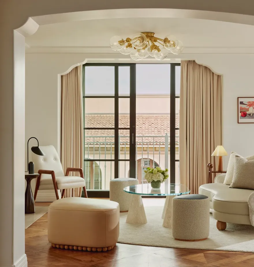 Intimate sitting room facing balcony doors; glass coffee table on conical bases, boucle stools, mid‑century chair and loveseat; terracotta roofline visible outside.