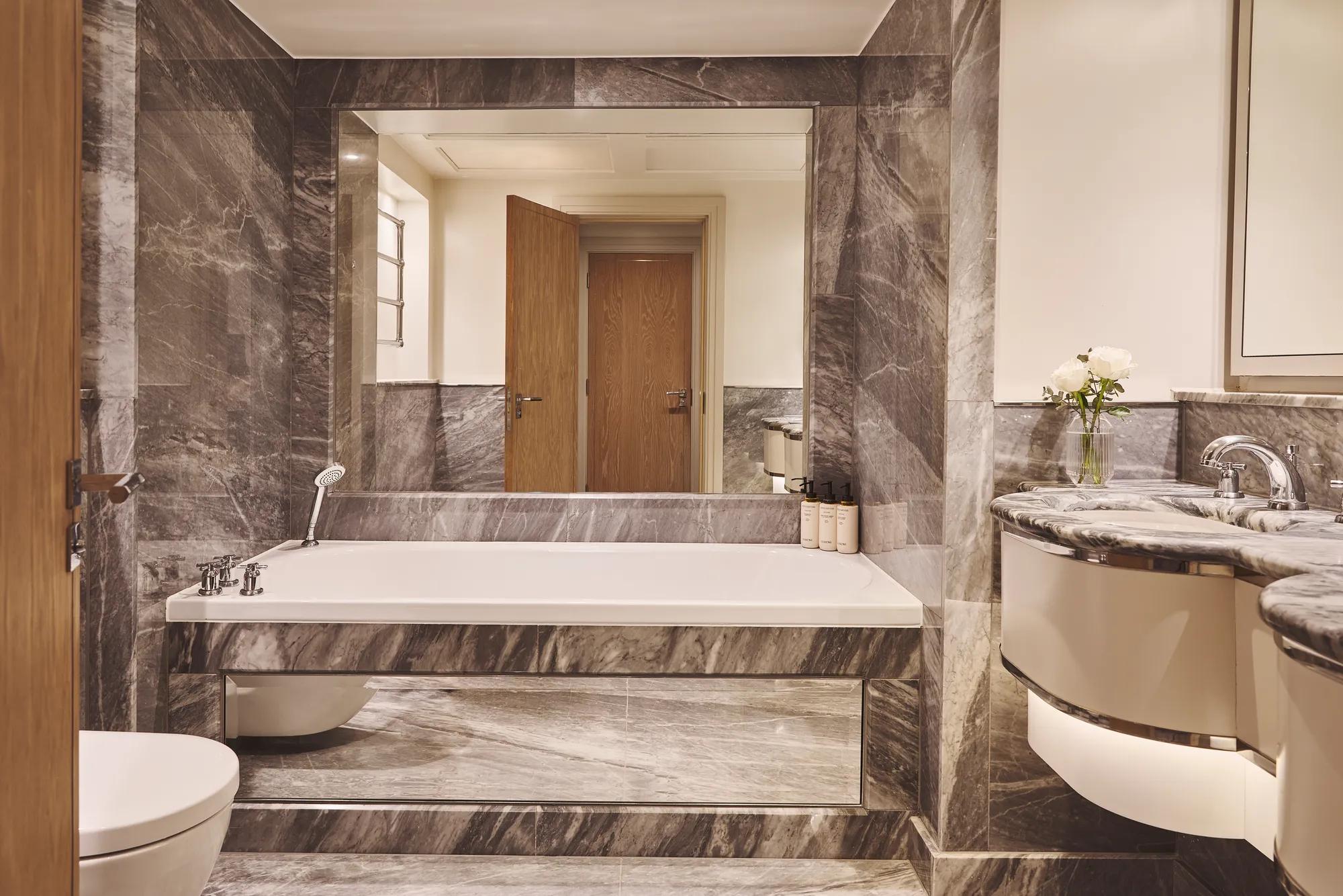 Marble bathroom with built-in bathtub, handheld shower, large mirror, floating vanity and wood door reflected in mirror.