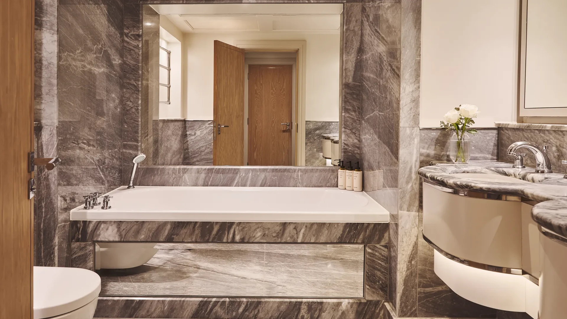 Marble bathroom with built-in bathtub, handheld shower, large mirror, floating vanity and wood door reflected in mirror.
