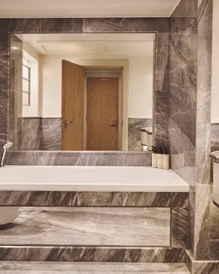 Marble bathroom with built-in bathtub, handheld shower, large mirror, floating vanity and wood door reflected in mirror.