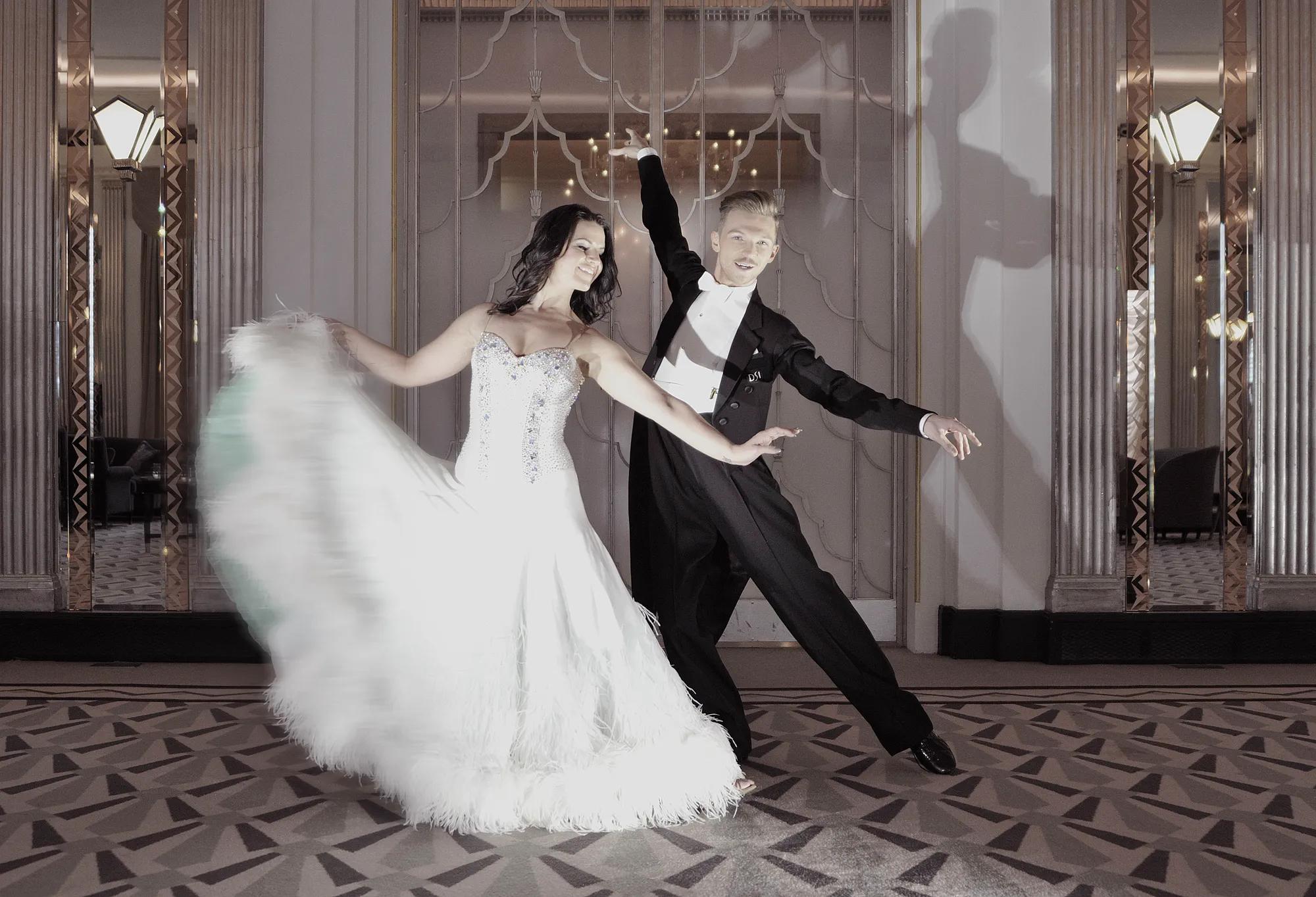 Elegant couple dancing in a grand ballroom, with the woman in a white feathered gown and the man in a black tailcoat, posing gracefully on a patterned floor beneath mirrored walls and soft lighting.