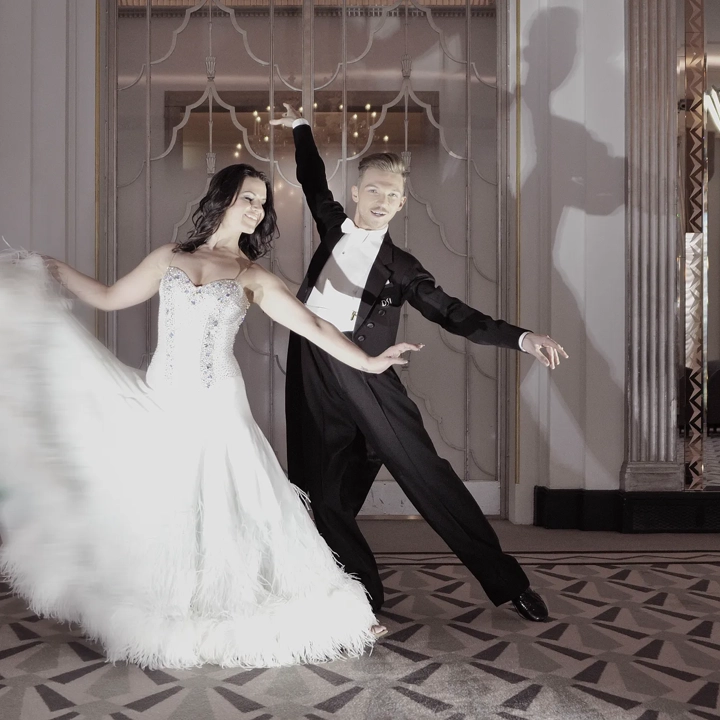 Elegant couple dancing in a grand ballroom, with the woman in a white feathered gown and the man in a black tailcoat, posing gracefully on a patterned floor beneath mirrored walls and soft lighting.