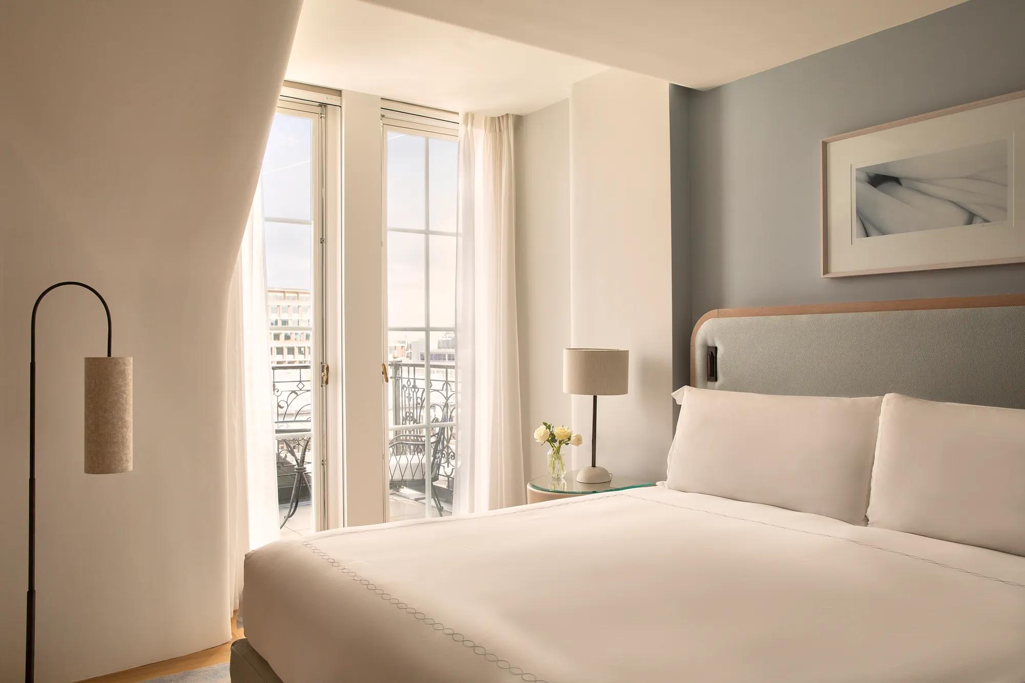 A bright and contemporary guest room featuring a king-size bed with crisp white linens, soft neutral furnishings and floor-to-ceiling French doors opening onto a private balcony. Natural light fills the space, highlighting the calm, understated design and city views beyond.