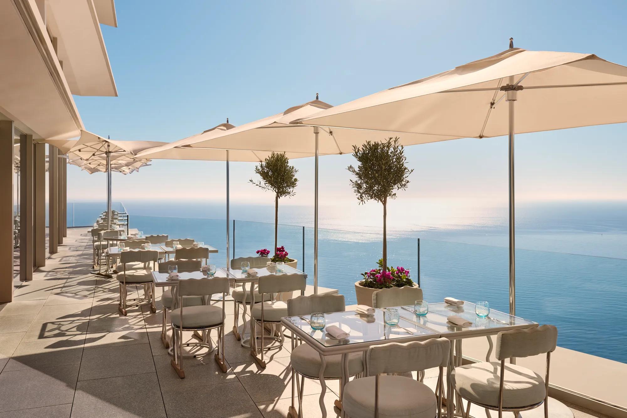 Terrasse avec tables dressées sous de grands parasols, offrant une vue sur la mer bleue et un ciel dégagé.