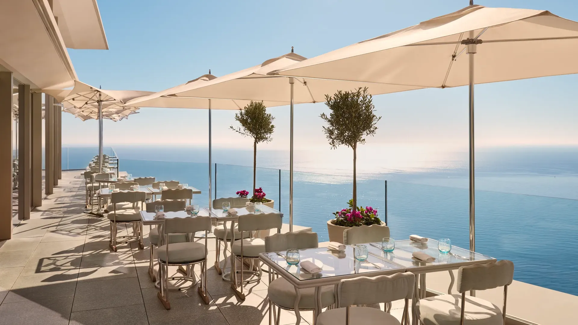 Terrasse avec tables dressées sous de grands parasols, offrant une vue sur la mer bleue et un ciel dégagé.