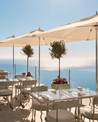 Terrasse avec tables dressées sous de grands parasols, offrant une vue sur la mer bleue et un ciel dégagé.