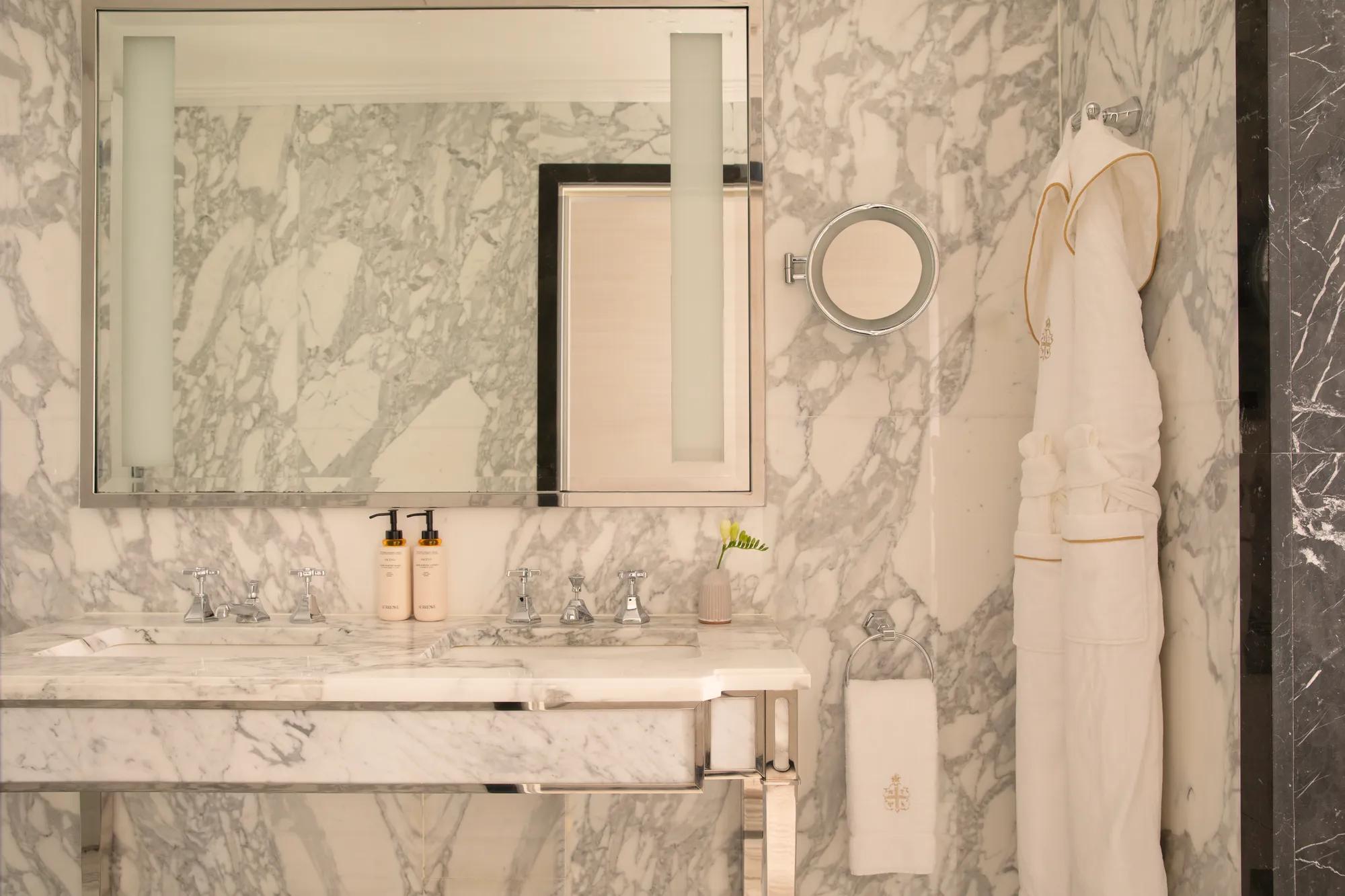 Marble bathroom with a double vanity, large mirror, luxury toiletries, white bathrobe and soft natural light.