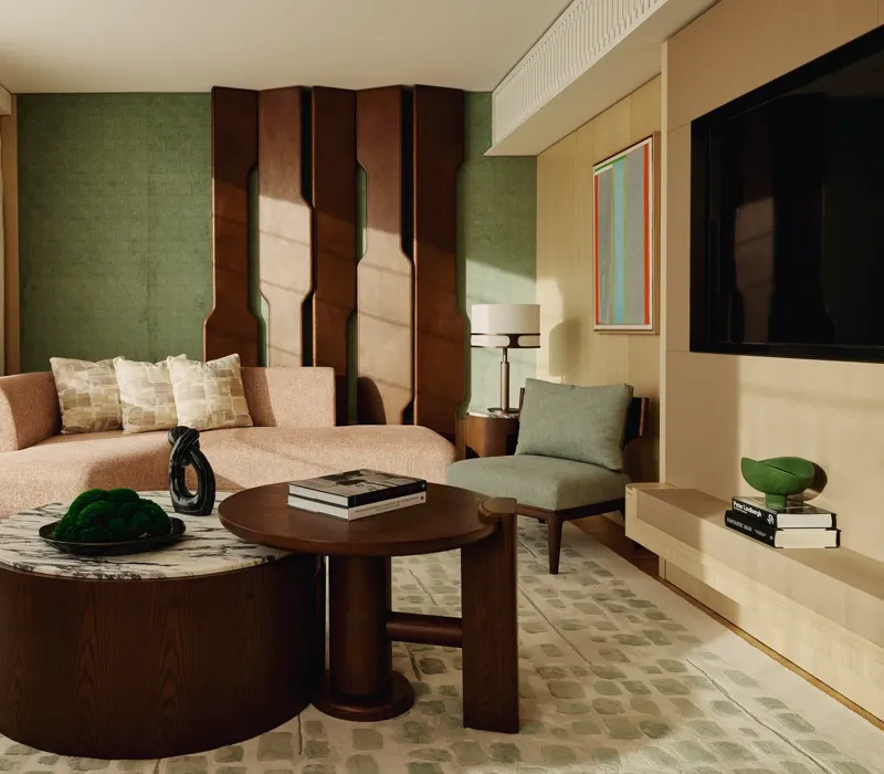 Stylish suite lounge with curved pink sofa, sculptural wall panel, and dual round coffee tables on green-patterned rug.