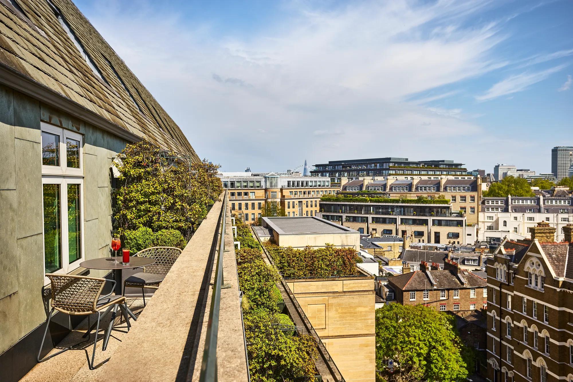 Balcony terrace with table and chairs overlooking rooftops, greenery, and a sunlit city skyline.