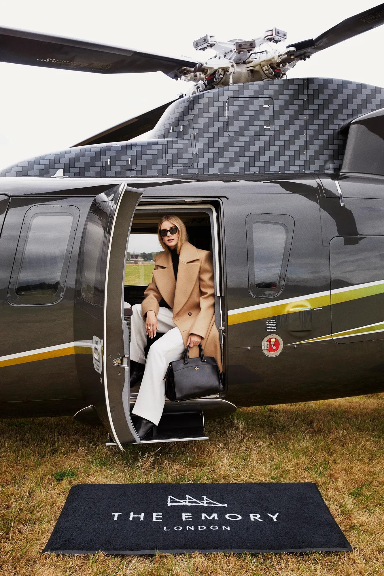 Guest steps off black helicopter onto branded mat reading “The Emory London” on a grass airfield.