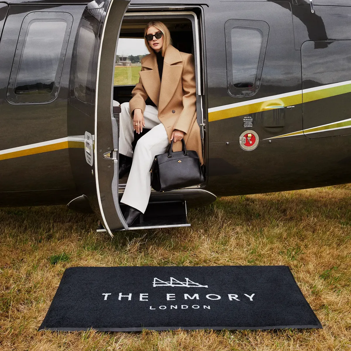 Guest steps off black helicopter onto branded mat reading “The Emory London” on a grass airfield.