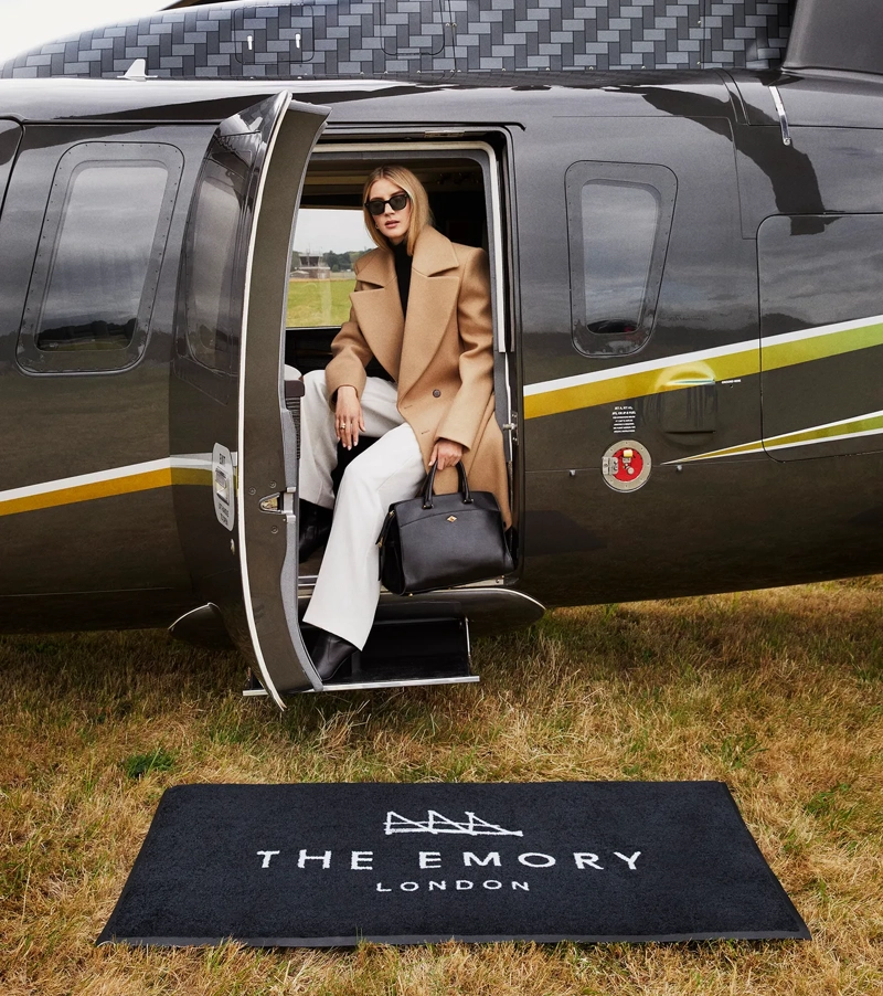 Guest steps off black helicopter onto branded mat reading “The Emory London” on a grass airfield.