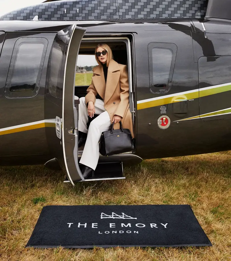 Guest steps off black helicopter onto branded mat reading “The Emory London” on a grass airfield.