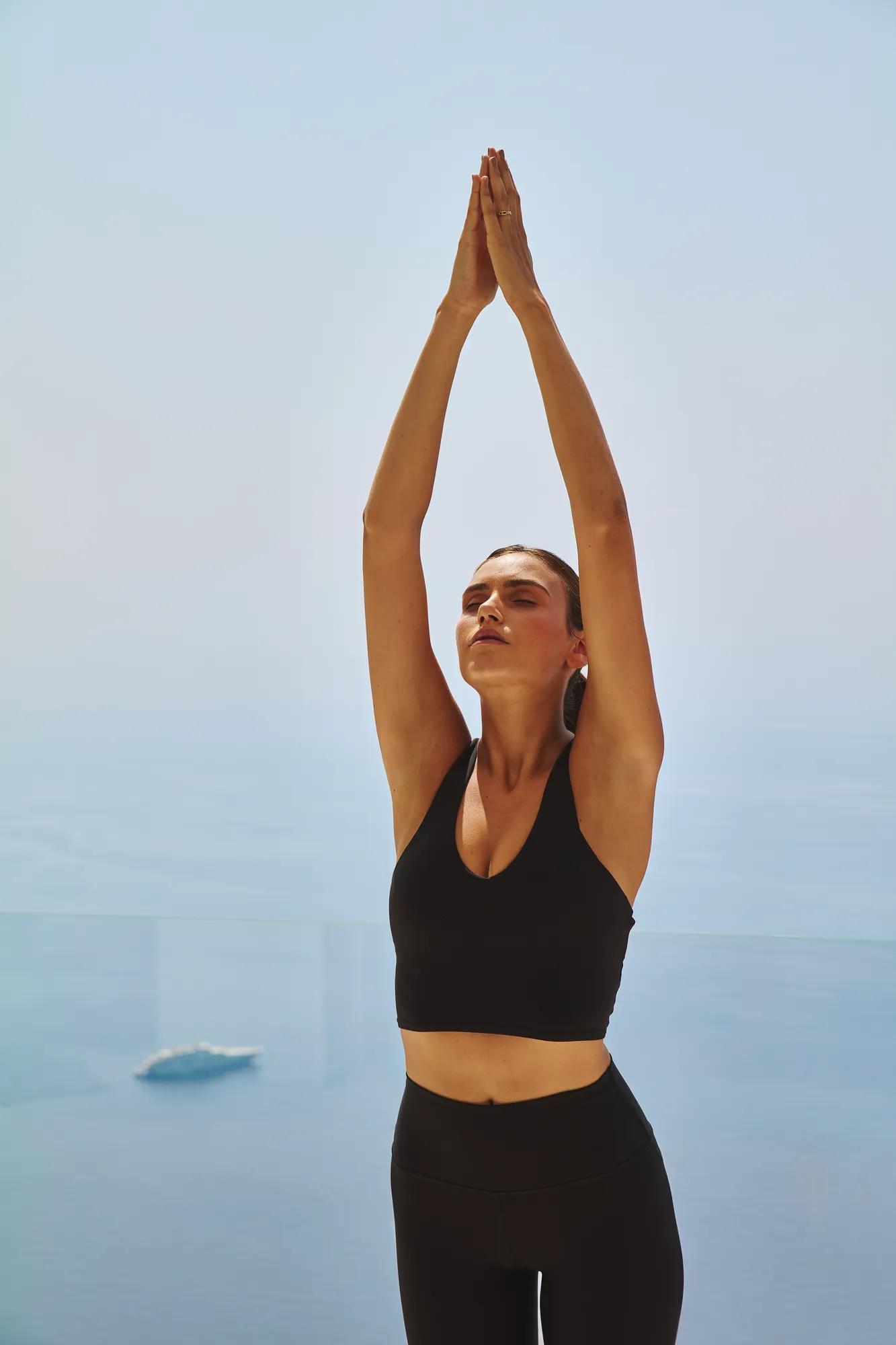 Femme en tenue de sport noire pratiquant le yoga, bras levés, sur une terrasse surplombant la mer avec un yacht au loin.