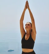 Femme en tenue de sport noire pratiquant le yoga, bras levés, sur une terrasse surplombant la mer avec un yacht au loin.