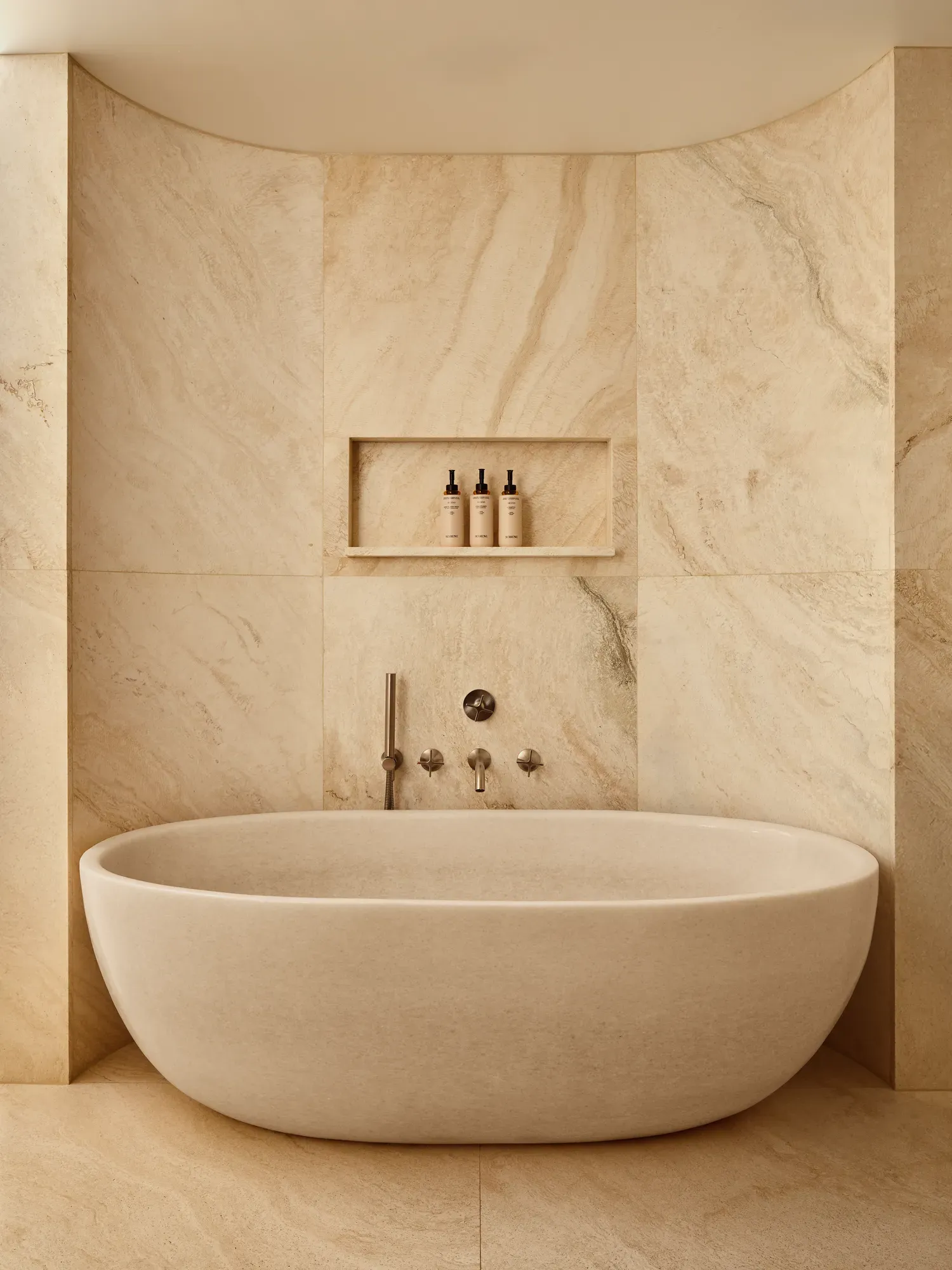 Spa-like bathroom with a sculpted freestanding stone bathtub, matching stone walls, and recessed shelving holding toiletries.