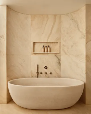Spa-like bathroom with a sculpted freestanding stone bathtub, matching stone walls, and recessed shelving holding toiletries.