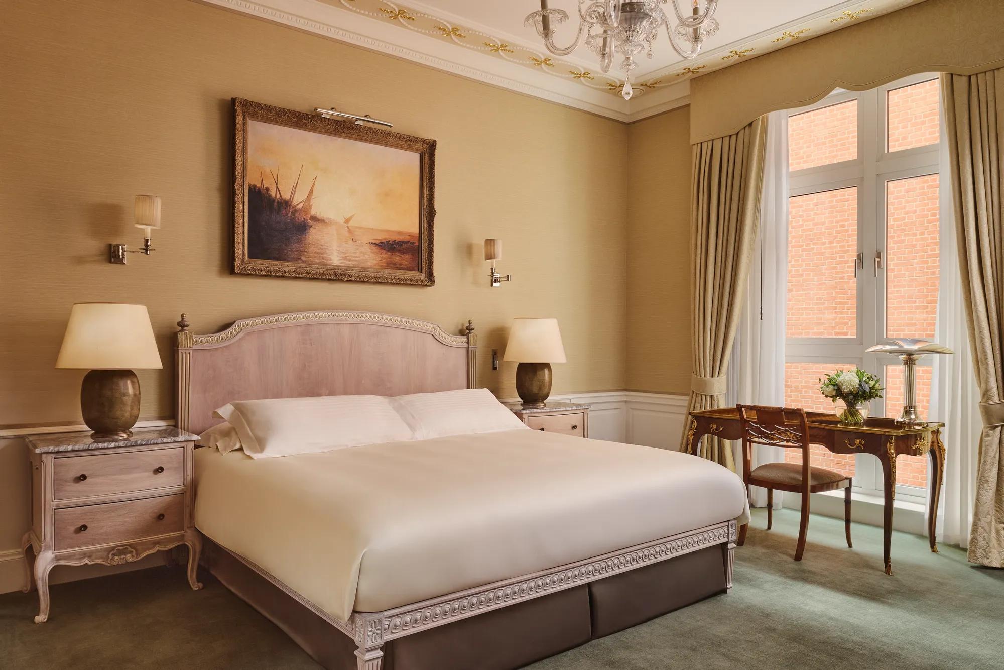 Classic hotel bedroom with king bed, vintage desk by window, and framed seascape painting.
