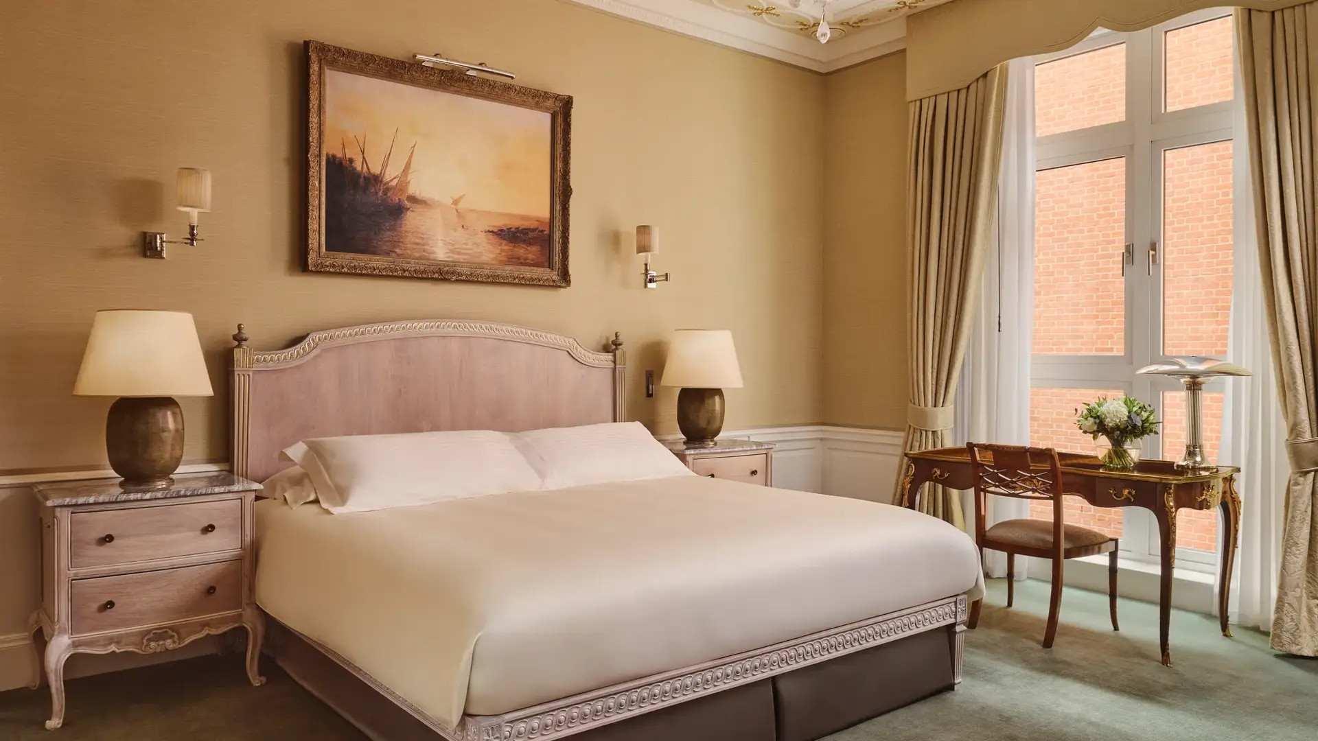 Classic hotel bedroom with king bed, vintage desk by window, and framed seascape painting.