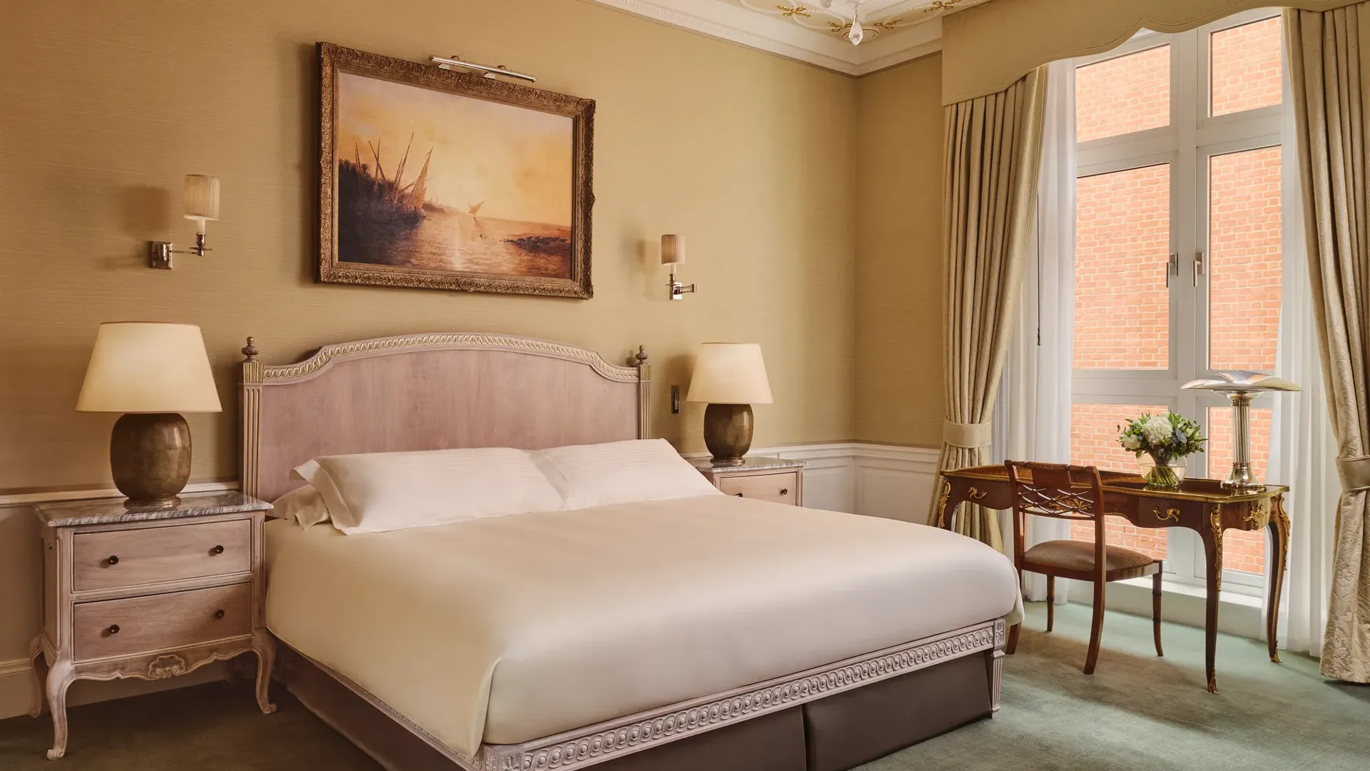 Classic hotel bedroom with king bed, vintage desk by window, and framed seascape painting.
