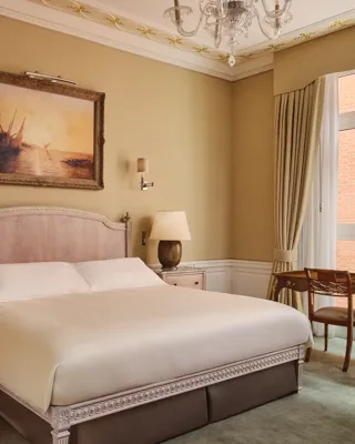 Classic hotel bedroom with king bed, vintage desk by window, and framed seascape painting.