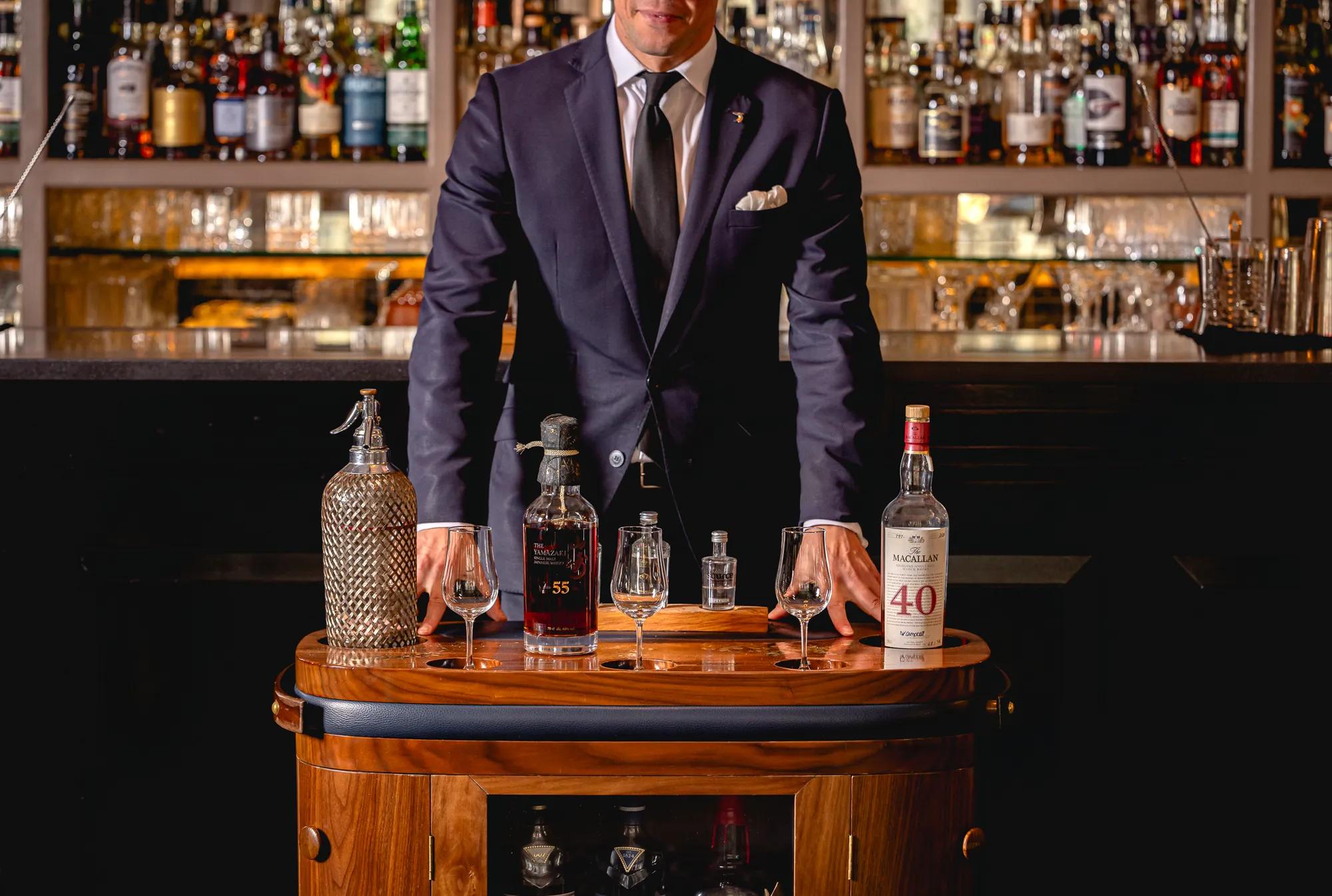 Server in a dark suit stands behind a drinks trolley displaying rare whisky bottles and tasting glasses in a bar.
