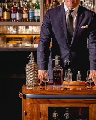 Server in a dark suit stands behind a drinks trolley displaying rare whisky bottles and tasting glasses in a bar.