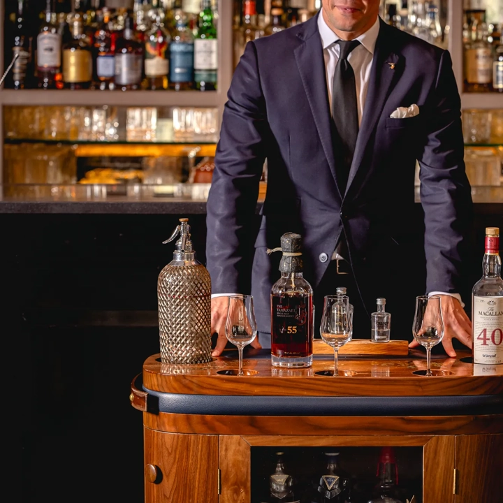 Server in a dark suit stands behind a drinks trolley displaying rare whisky bottles and tasting glasses in a bar.