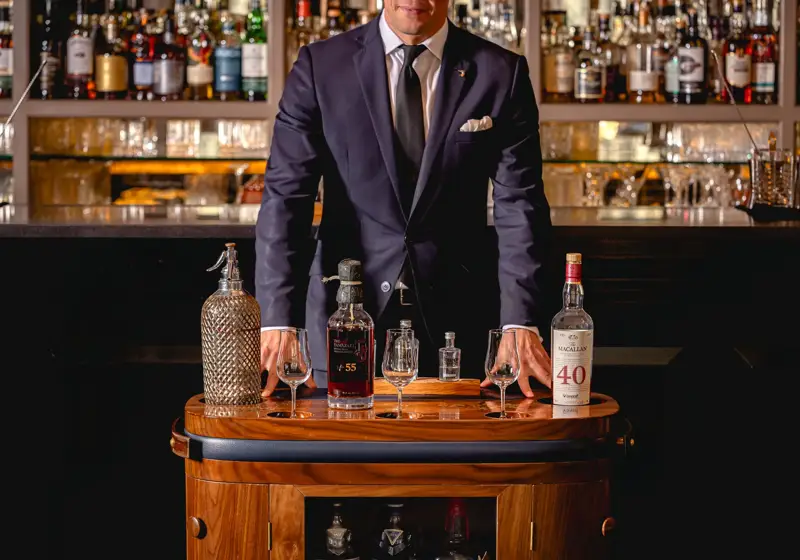 Server in a dark suit stands behind a drinks trolley displaying rare whisky bottles and tasting glasses in a bar.
