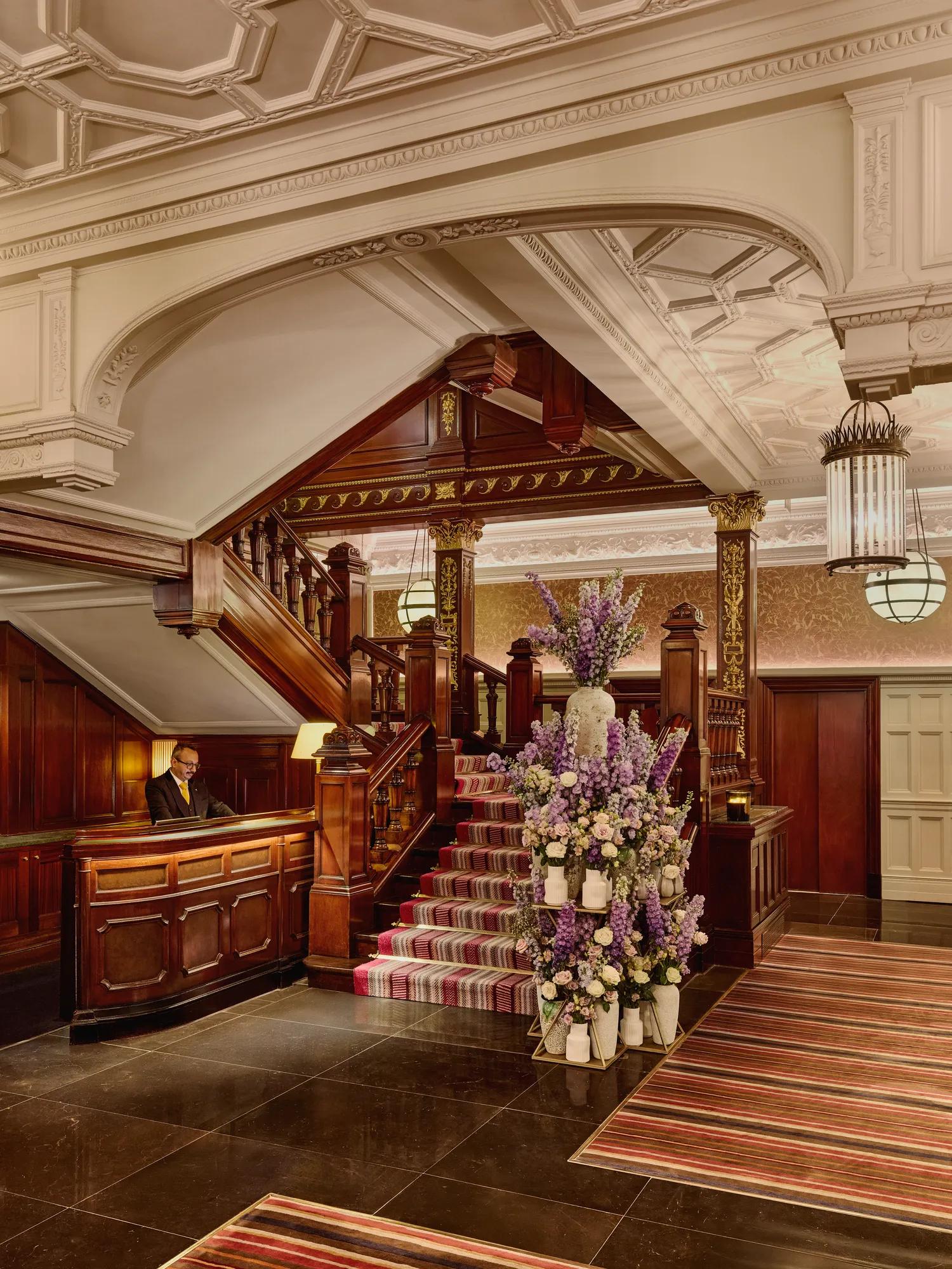 Elegant hotel lobby with dark wood staircase, ornate ceiling, striped carpet, and large floral arrangement near the reception desk.
