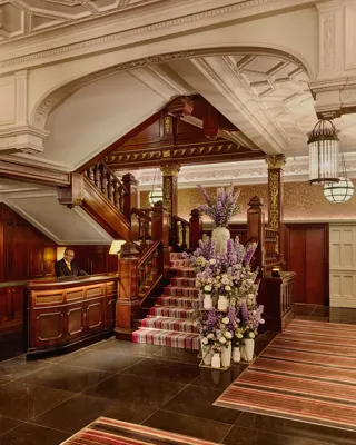 Elegant hotel lobby with dark wood staircase, ornate ceiling, striped carpet, and large floral arrangement near the reception desk.