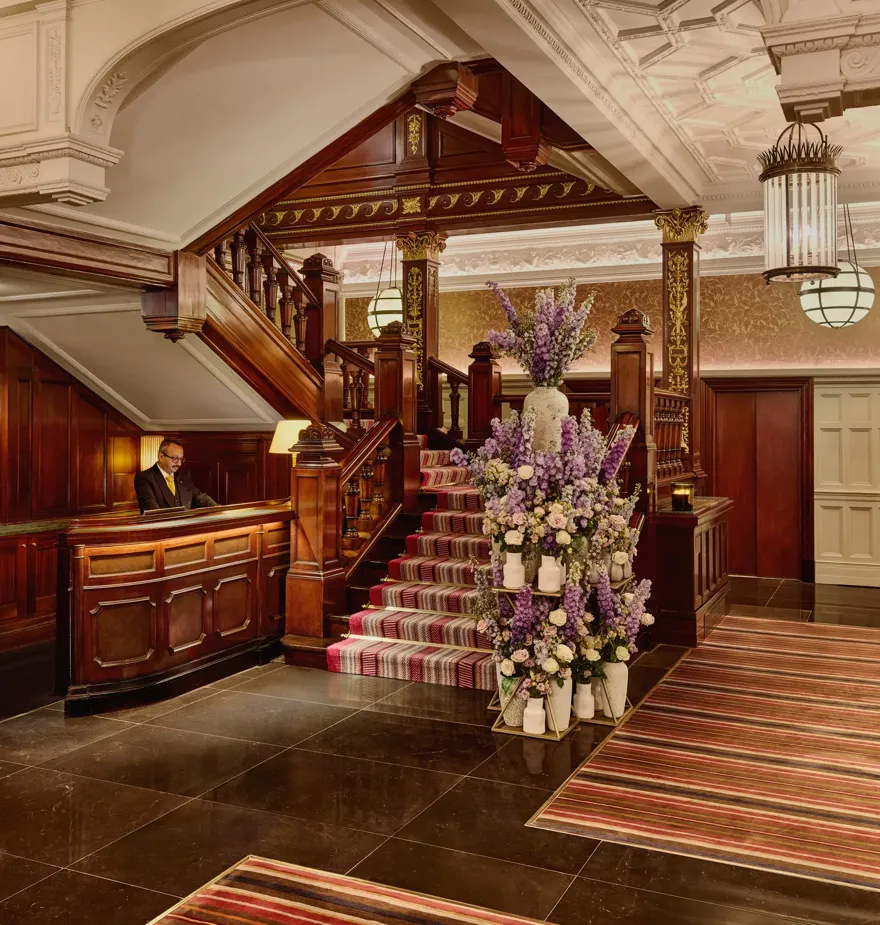 Elegant hotel lobby with dark wood staircase, ornate ceiling, striped carpet, and large floral arrangement near the reception desk.