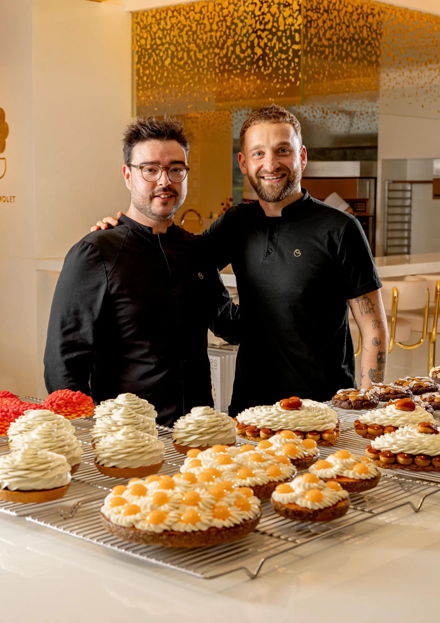 Two pastry chefs stand behind trays of fruit tarts and cream-topped pastries in a bright patisserie kitchen.