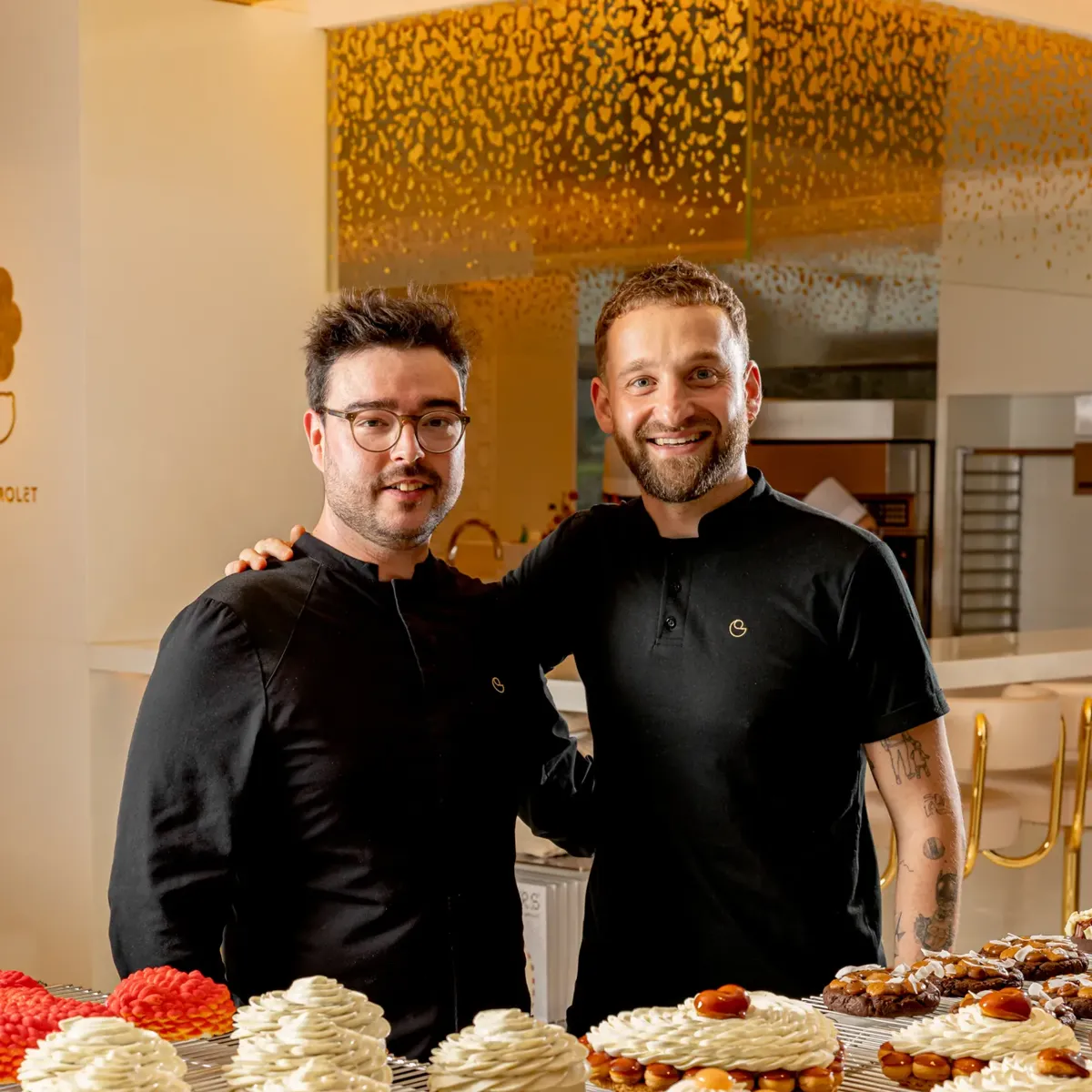 Two pastry chefs stand behind trays of fruit tarts and cream-topped pastries in a bright patisserie kitchen.