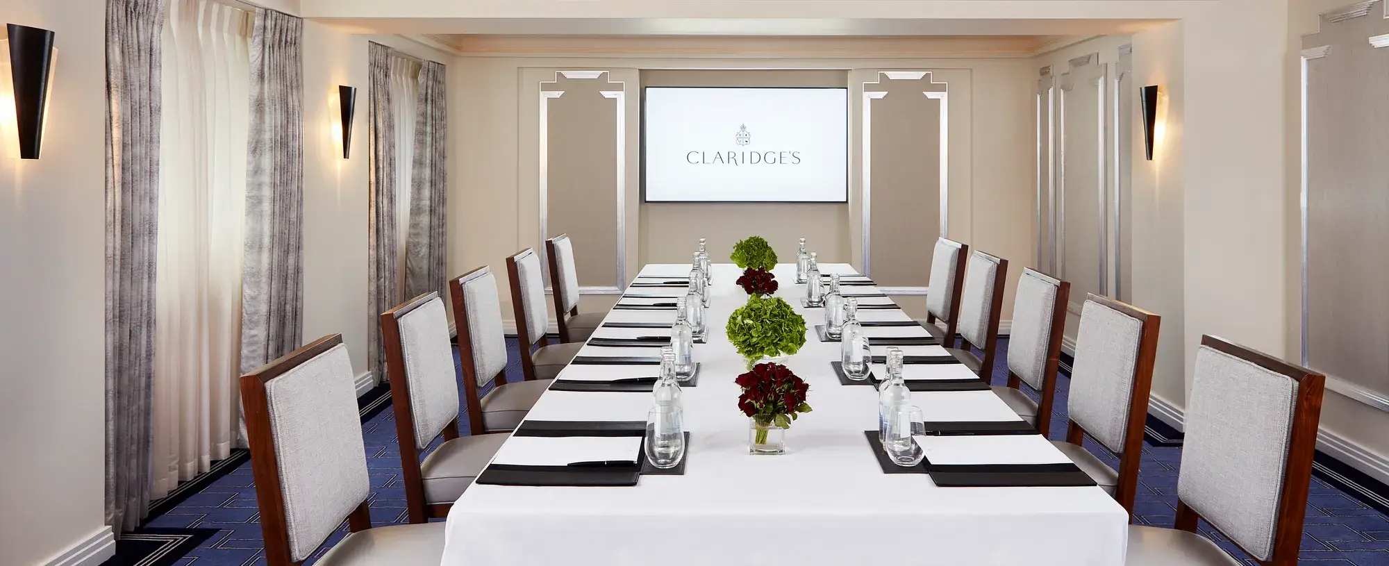 Stylish boardroom at Claridge’s with a long white-clothed table set for a meeting, surrounded by upholstered chairs, floral centrepieces, and a presentation screen at the far end.