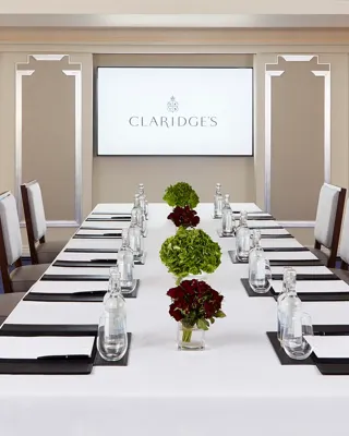 Stylish boardroom at Claridge’s with a long white-clothed table set for a meeting, surrounded by upholstered chairs, floral centrepieces, and a presentation screen at the far end.