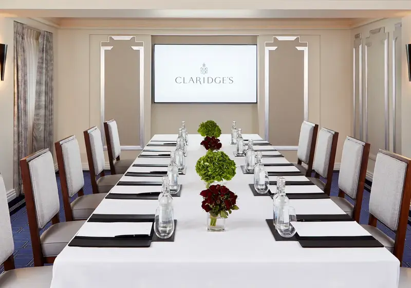 Stylish boardroom at Claridge’s with a long white-clothed table set for a meeting, surrounded by upholstered chairs, floral centrepieces, and a presentation screen at the far end.