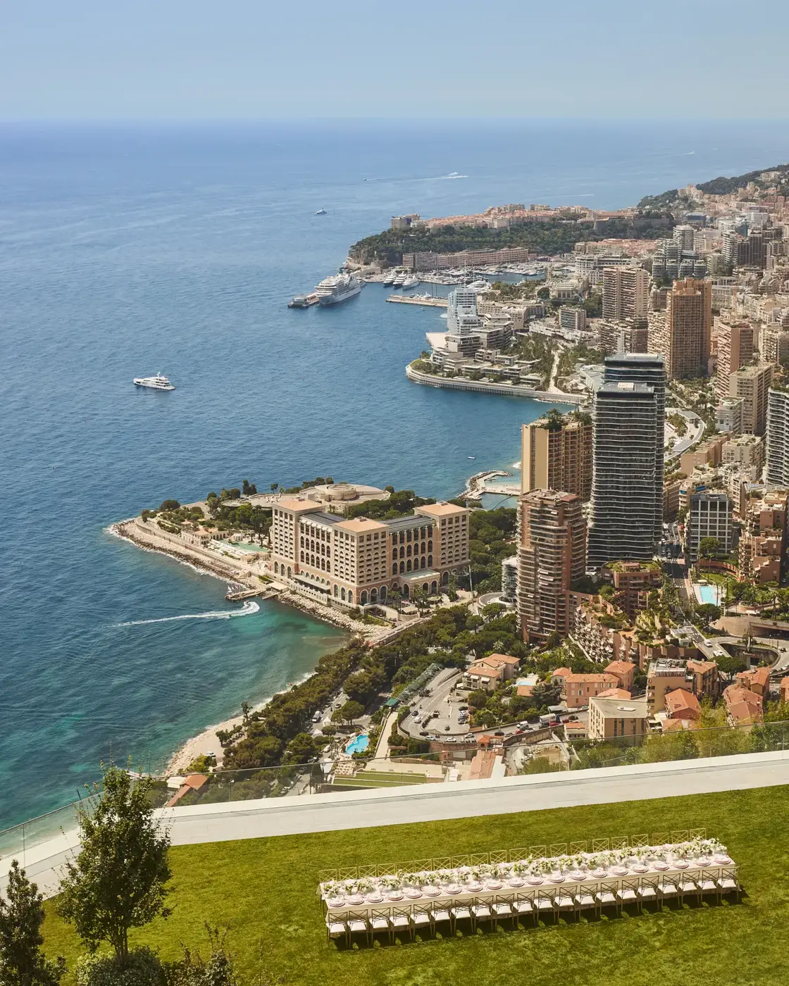 Vue panoramique du littoral de Monaco avec immeubles, yachts en mer et terrasse herbeuse au premier plan.