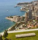 Vue panoramique du littoral de Monaco avec immeubles, yachts en mer et terrasse herbeuse au premier plan.