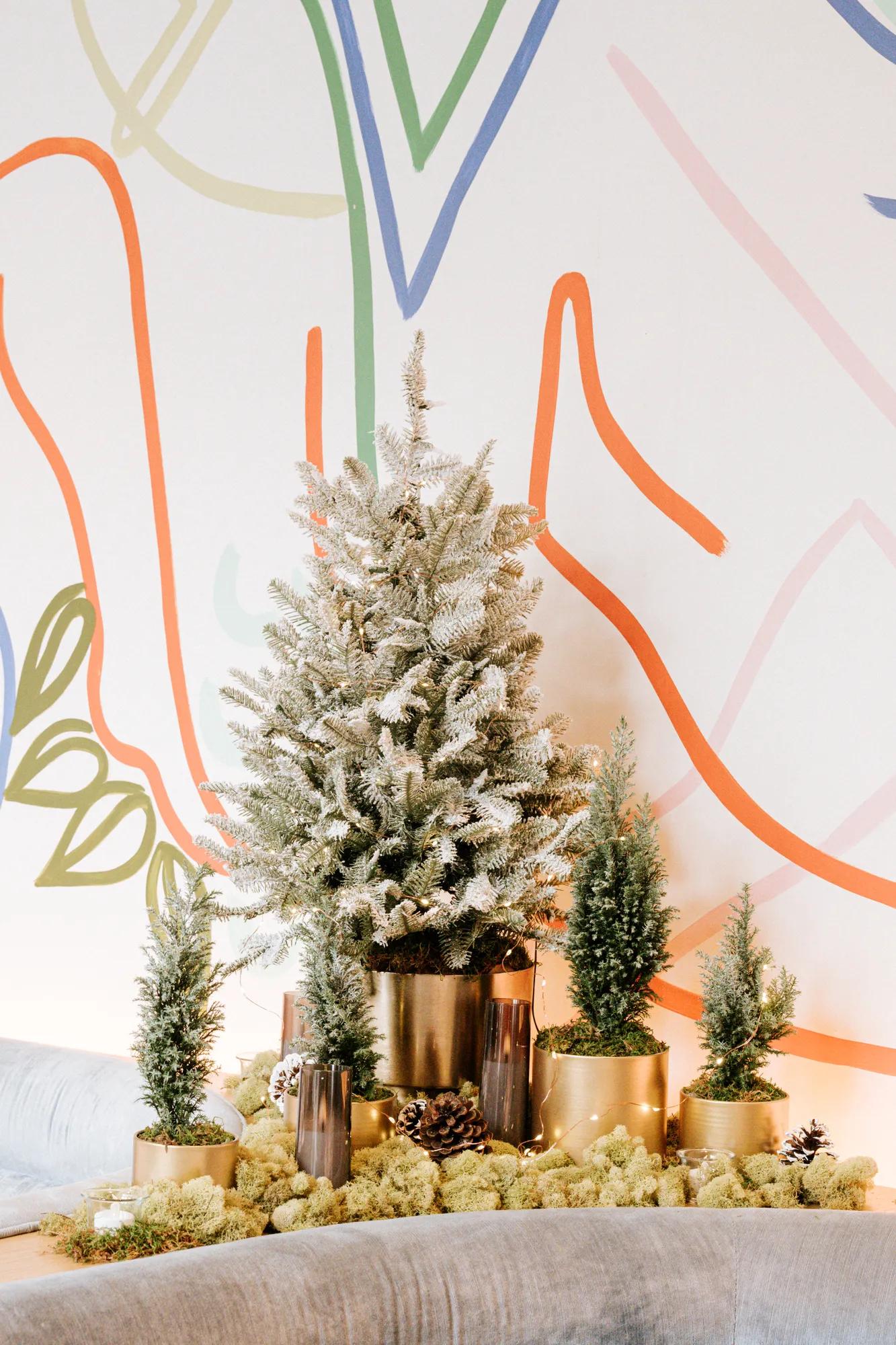 Festive evergreen tree display with frosted foliage, candlelight accents, and gold planters set against a colorful mural backdrop.