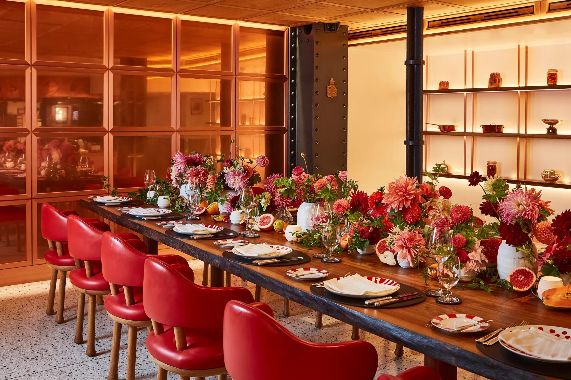 Vibrant private dining room at Claridge’s with a long wooden table set with red chairs, striped plates, and abundant floral centrepieces in red and pink hues.