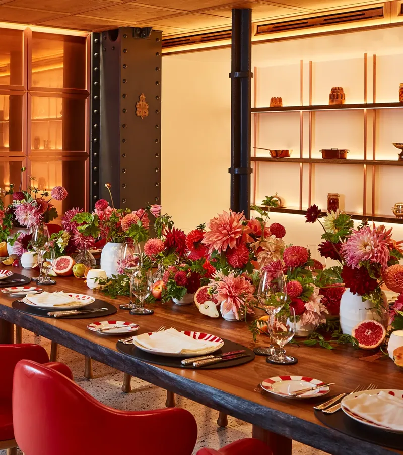 Vibrant private dining room at Claridge’s with a long wooden table set with red chairs, striped plates, and abundant floral centrepieces in red and pink hues.