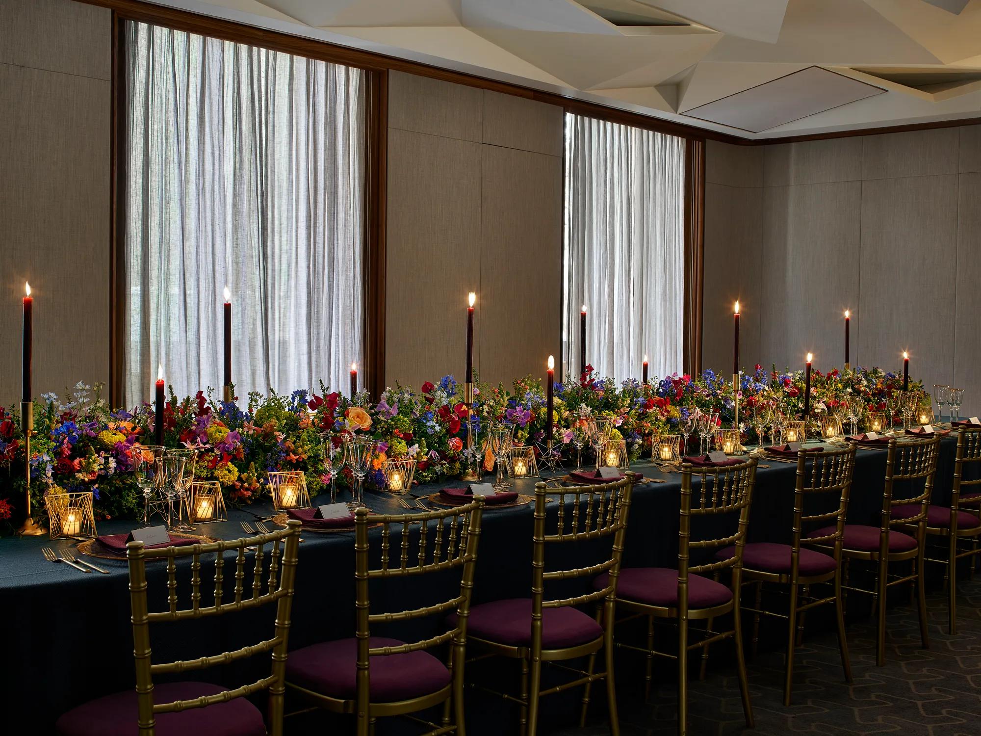 Long banquet table with gold chairs, dark tablecloth, colourful flowers, and tall candles against curtained windows