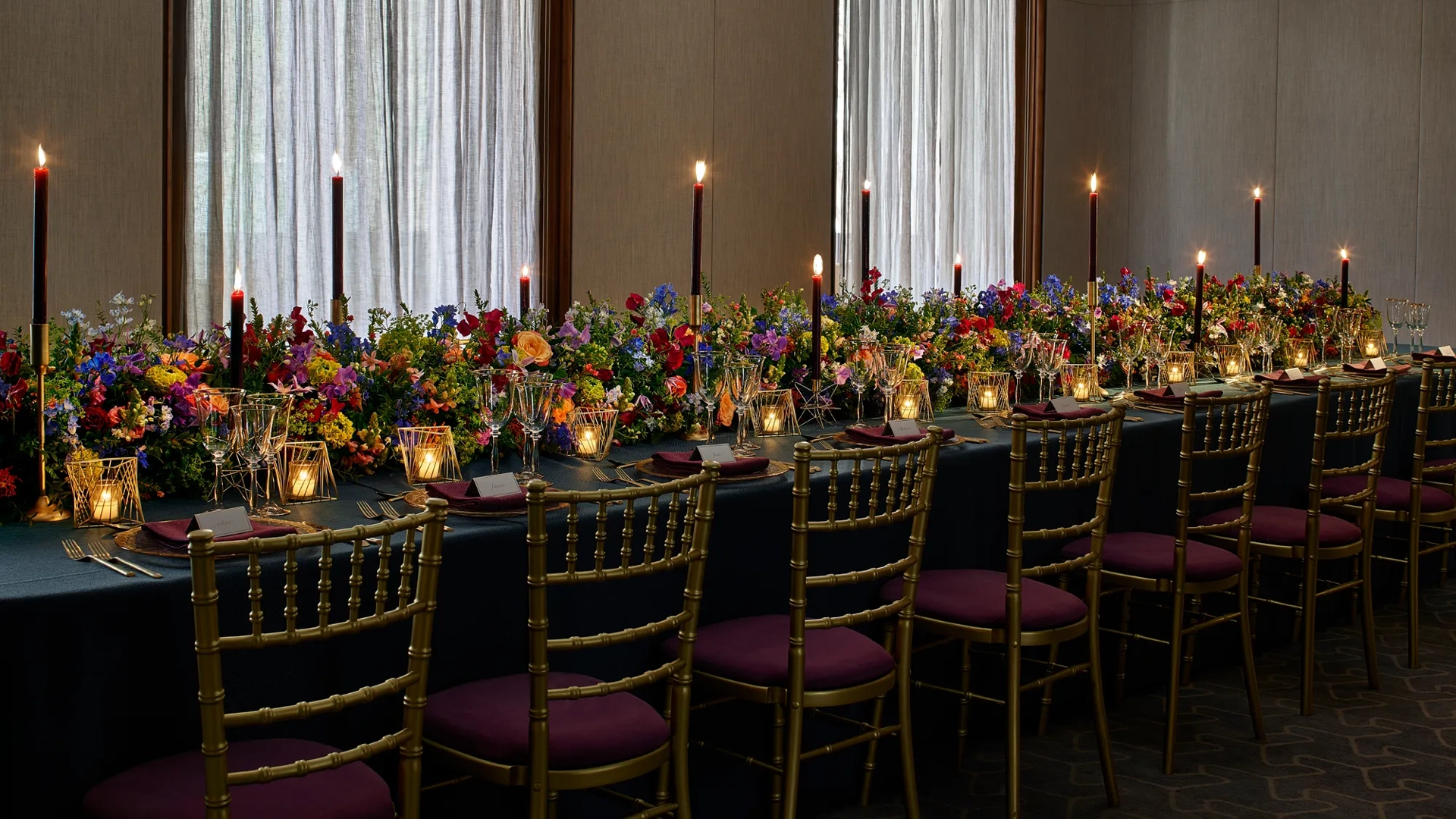 Long banquet table with gold chairs, dark tablecloth, colourful flowers, and tall candles against curtained windows