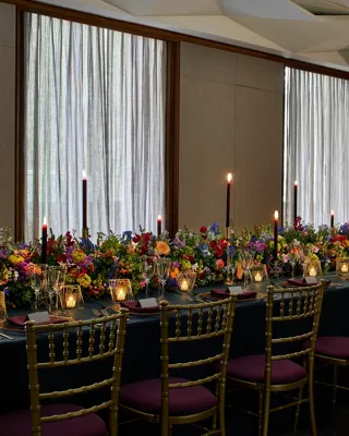 Long banquet table with gold chairs, dark tablecloth, colourful flowers, and tall candles against curtained windows