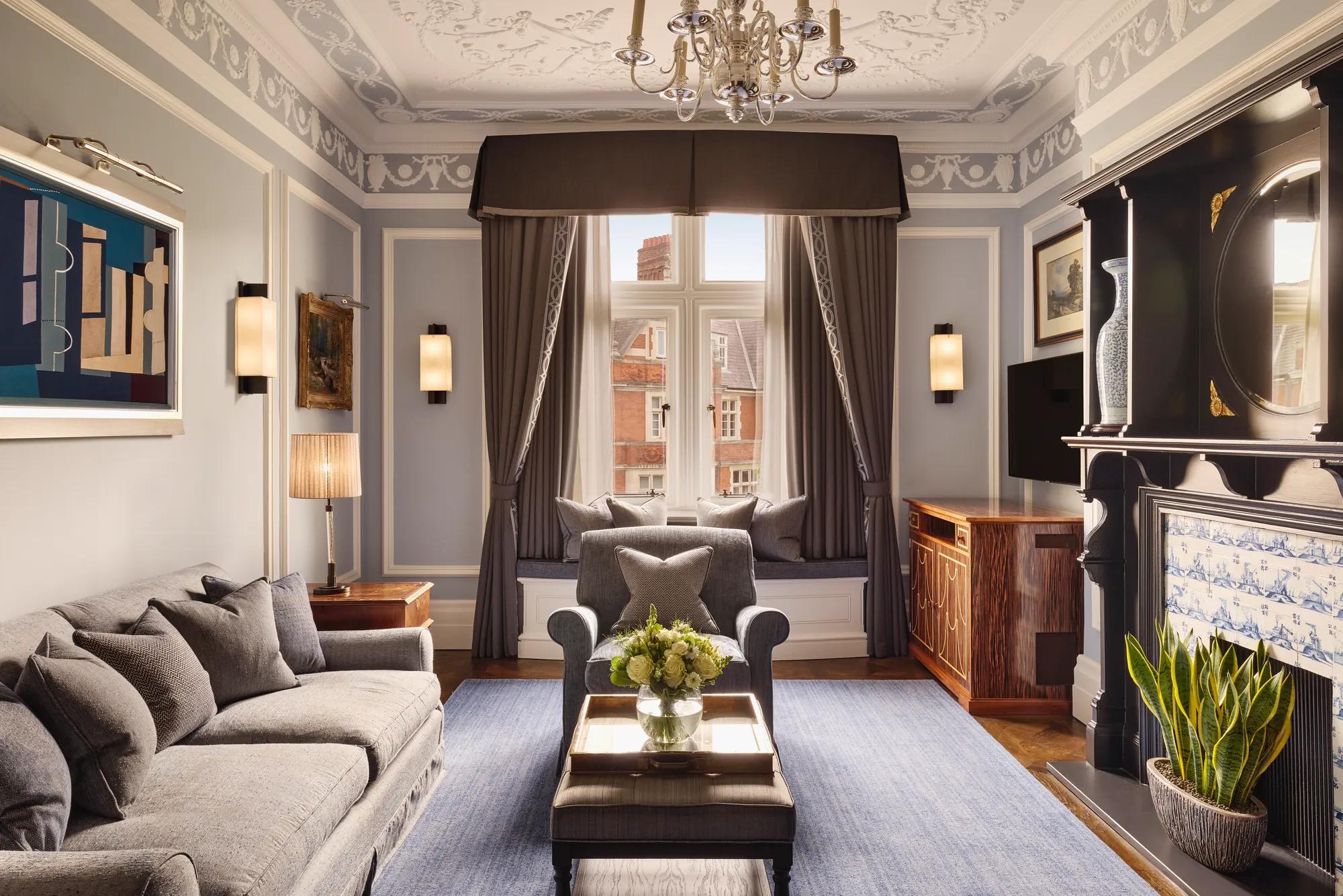 Elegant living room with grey sofa and armchairs, fireplace, window seat with cushions, and large window with drapes.