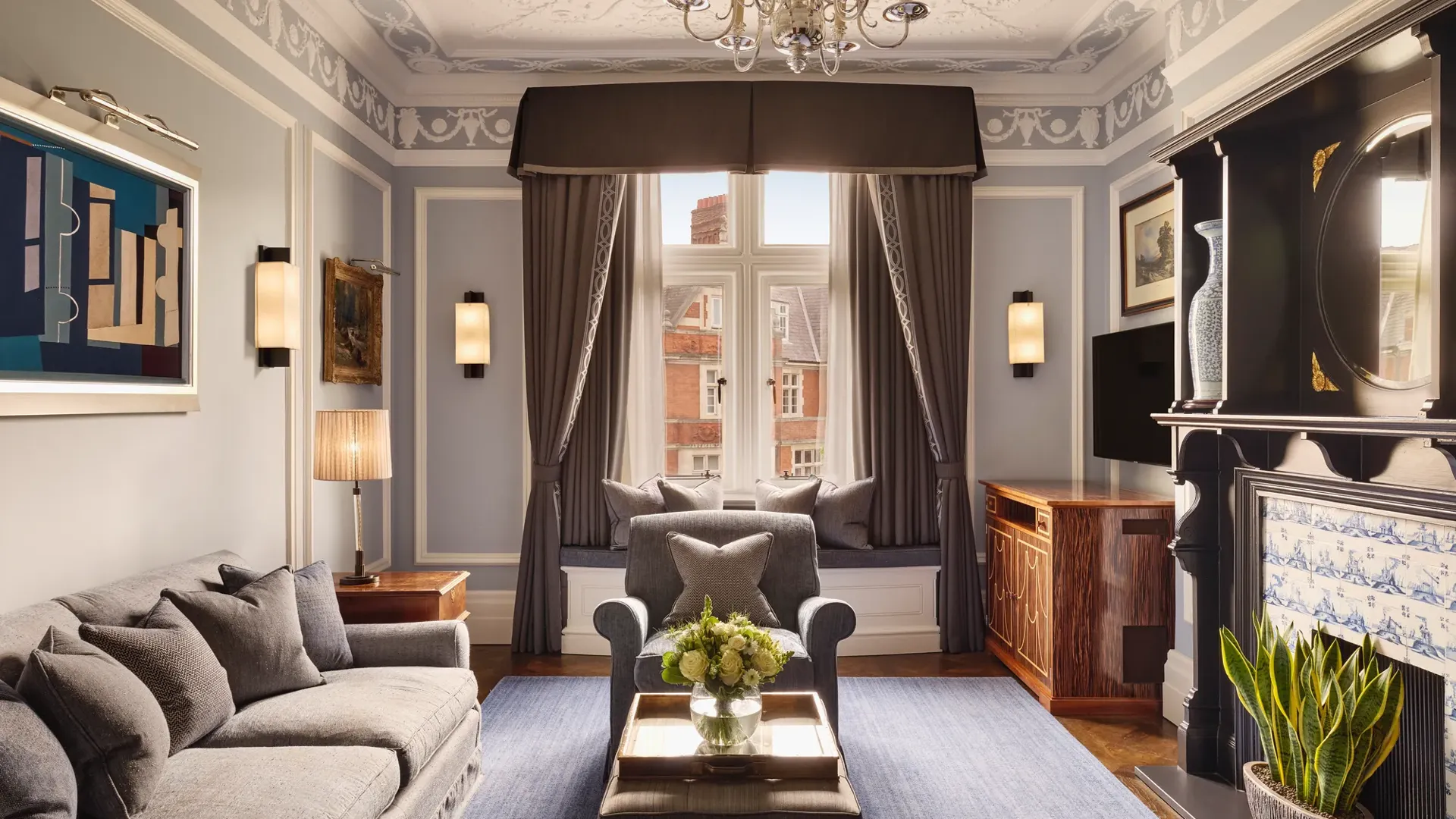 Elegant living room with grey sofa and armchairs, fireplace, window seat with cushions, and large window with drapes.