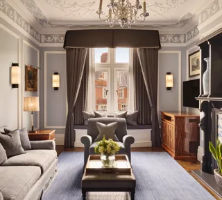 Elegant living room with grey sofa and armchairs, fireplace, window seat with cushions, and large window with drapes.