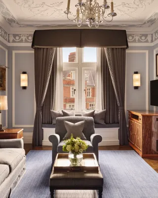Elegant living room with grey sofa and armchairs, fireplace, window seat with cushions, and large window with drapes.